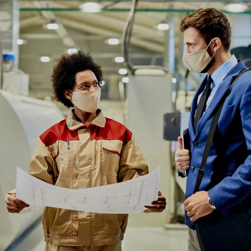 African American female worker and businessman wearing face masks while examining blueprints at woodworking production facility.