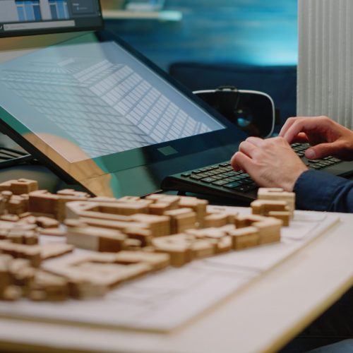 Close up of architect hands using touch screen computer and maquette on table. Man engineer working with technology to analyze building model for construction layout and development