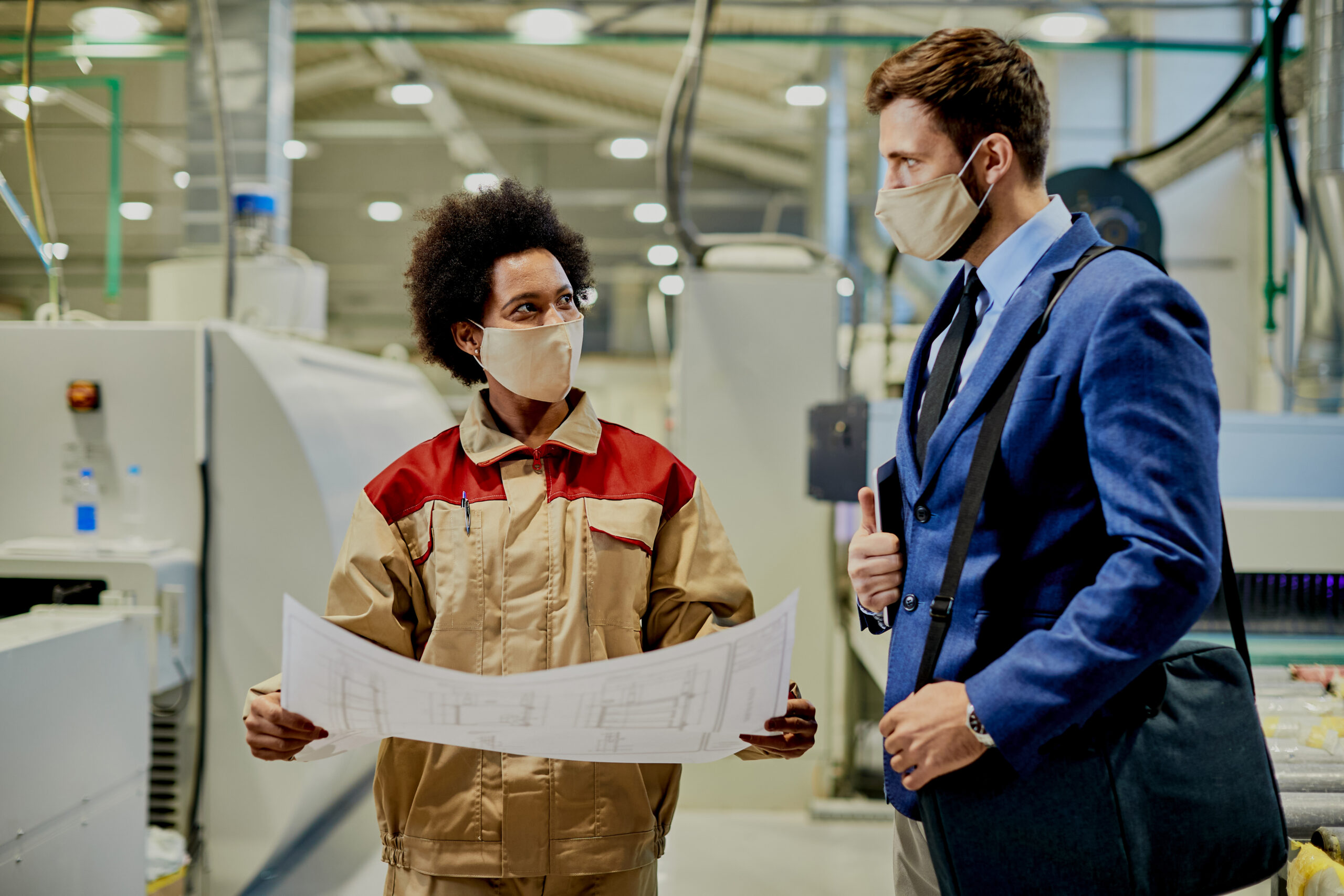 African American female worker and businessman wearing face masks while examining blueprints at woodworking production facility.